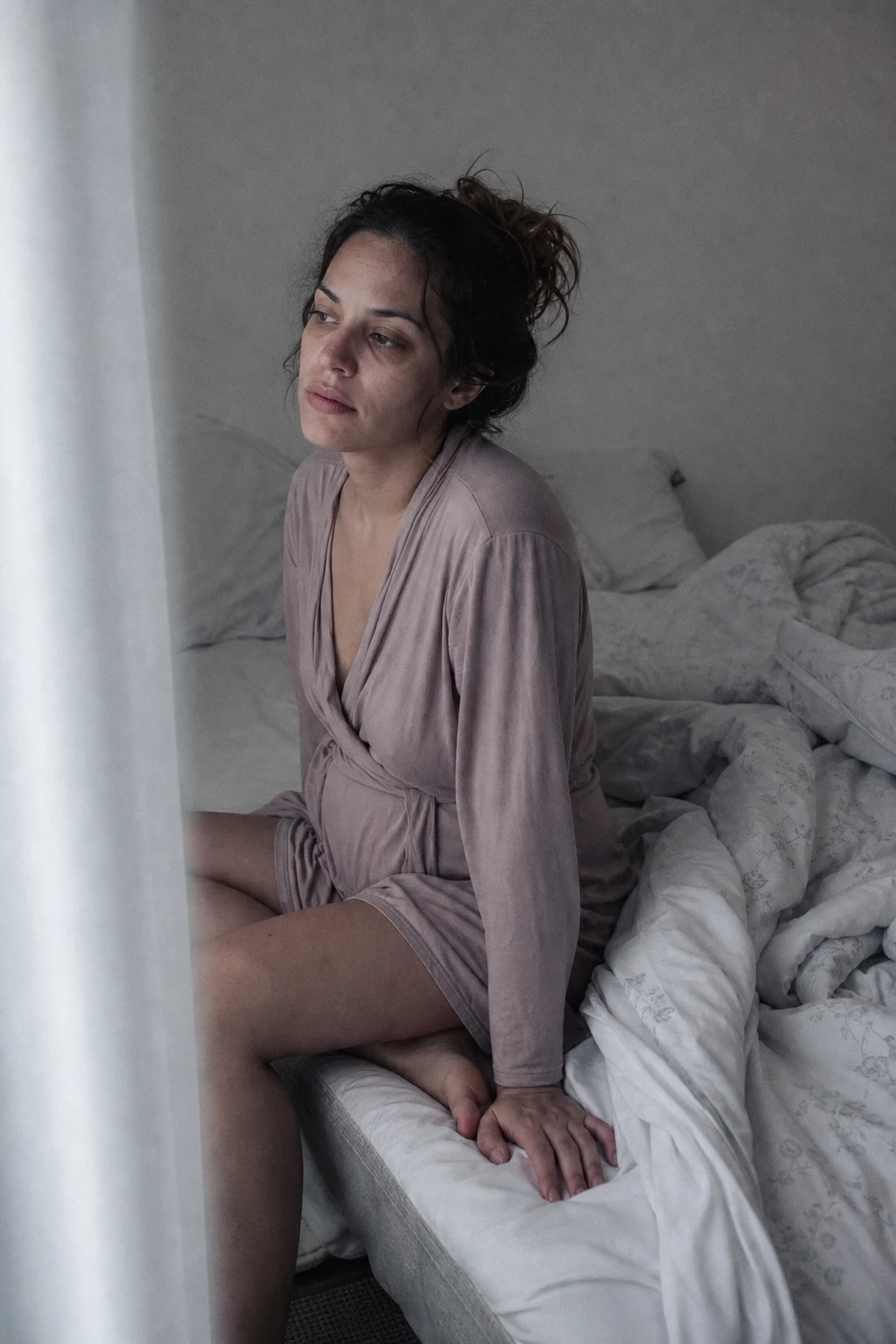 Mulher no pós-parto, tranquila e pensativa, em quarto de luz suave e granulada, com um tecido aconchegante por perto — íntima e humana